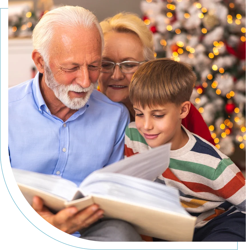 Elderly couple reading a book with a young boy by a decorated Christmas tree.